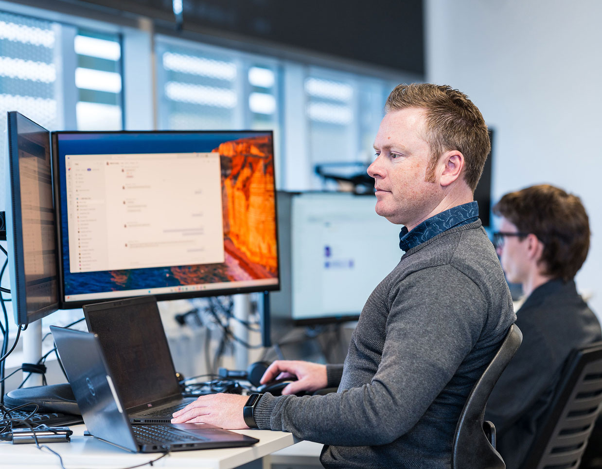 A man in a grey sweater works intently at a computer with dual monitors displaying software. Another person is in the background. The office setting is bright and modern.