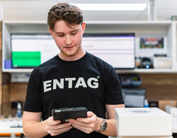 A young man with wavy hair, wearing a black "ENTAG" T-shirt, looks at a tablet. Shelves with electronics are in the blurred background, creating a focused, tech-savvy atmosphere.
