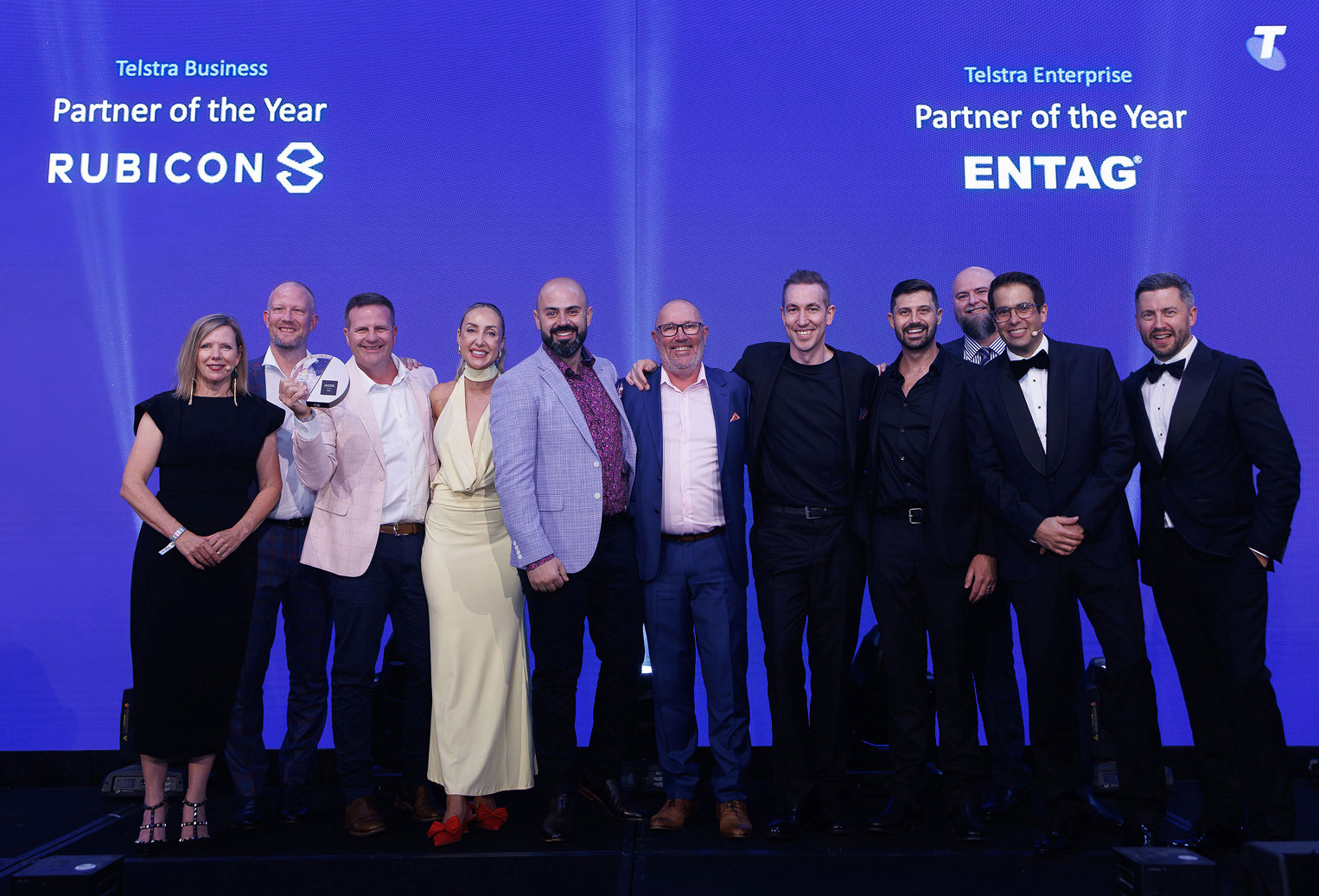 A group of ten people stands smiling on stage in formal attire. Behind them, a blue screen displays "Telstra Business Partner of the Year" awards.