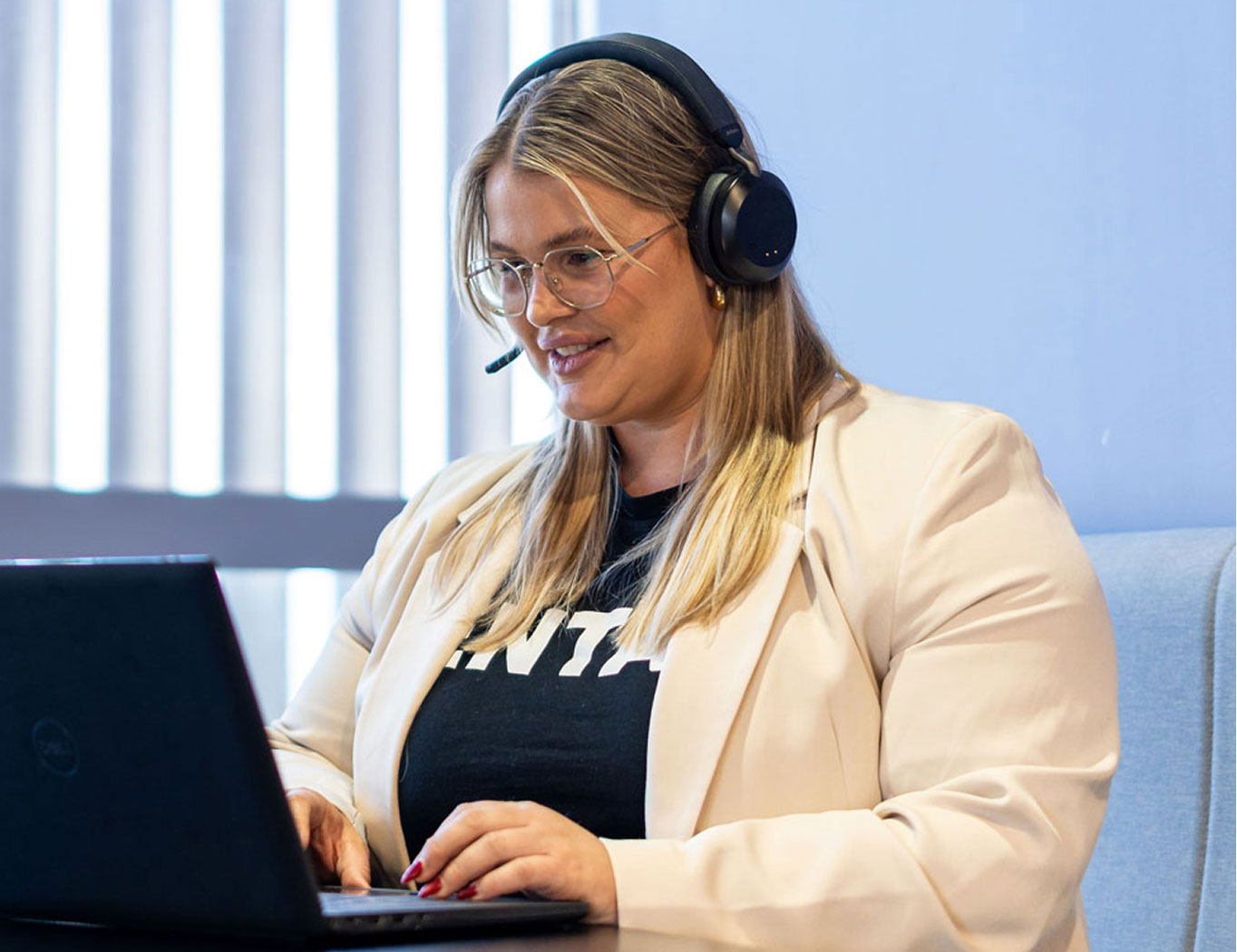 A woman wearing a headset and glasses smiles while working on a laptop. She is seated indoors with vertical blinds in the background, conveying a professional and focused atmosphere.